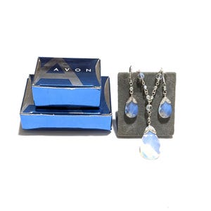 May include: A silver-toned jewelry set featuring a necklace and earrings with teardrop-shaped, iridescent blue pendants. The set is displayed on a gray velvet surface, next to two blue boxes with the word "AVON" printed on them.