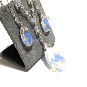 May include: A silver-toned jewelry set featuring a pair of drop earrings and a matching pendant necklace. Each piece showcases a large, pear-shaped, opalescent stone, accented with smaller clear crystals and delicate chain details. The set is displayed on a gray velvet stand.