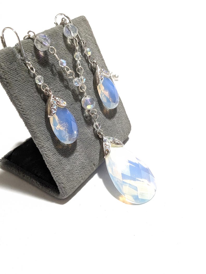 May include: A silver-toned jewelry set featuring a pair of drop earrings and a matching pendant necklace. Each piece showcases a large, pear-shaped, opalescent stone, accented with smaller clear crystals and delicate chain details. The set is displayed on a gray velvet stand.