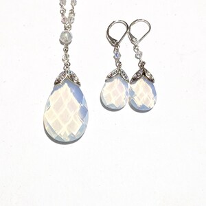 May include: A silver necklace and matching earrings set. The necklace features a teardrop-shaped pendant with a faceted, opalescent stone, accented with small crystals. The earrings mirror the pendant design, with lever-back closures. The jewelry set is displayed on a white background.