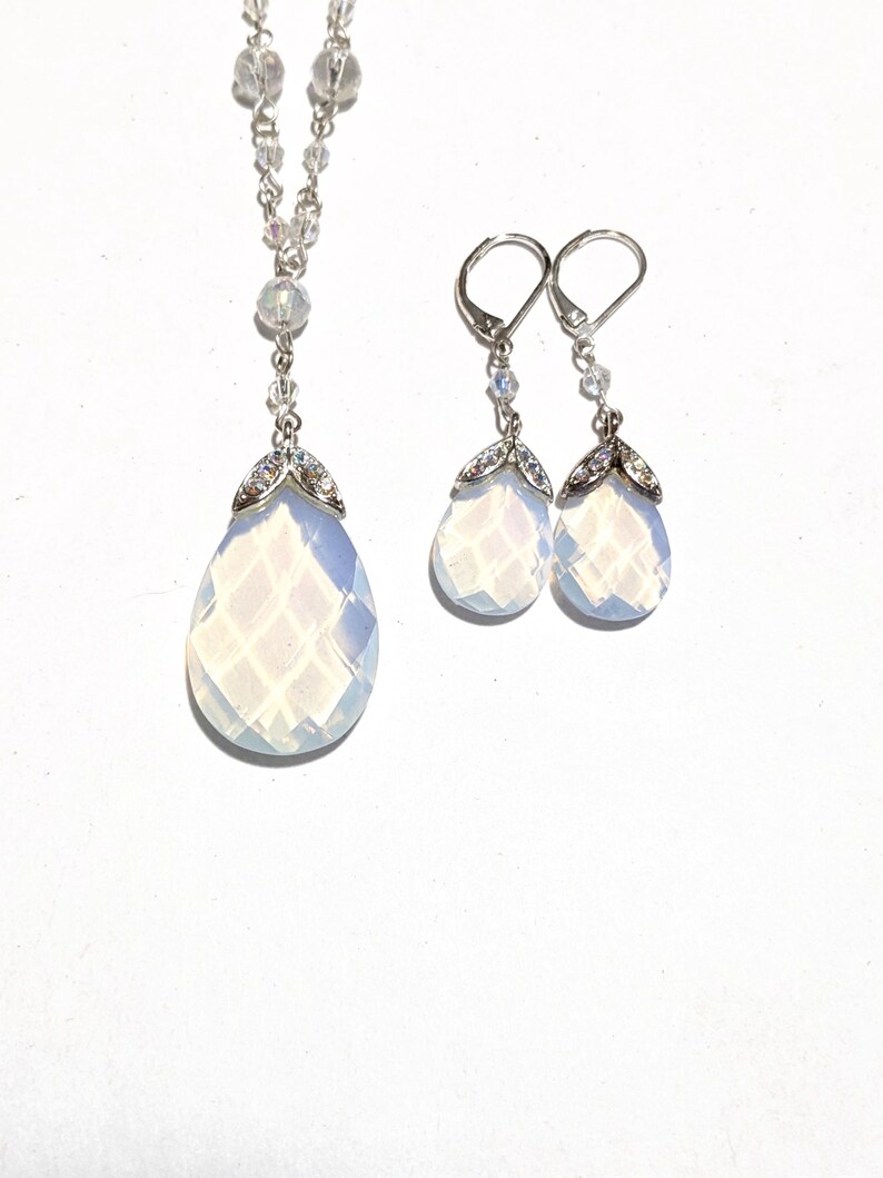May include: A silver necklace and matching earrings set. The necklace features a teardrop-shaped pendant with a faceted, opalescent stone, accented with small crystals. The earrings mirror the pendant design, with lever-back closures. The jewelry set is displayed on a white background.