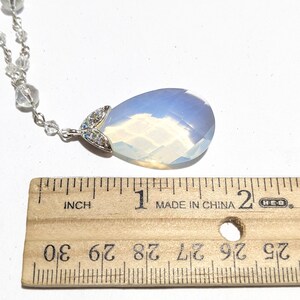 May include: A teardrop-shaped pendant necklace with a large, opalescent stone. The pendant is approximately 1 inch long and is attached to a delicate silver chain with small, clear beads. The pendant has a silver accent with small, clear stones.
