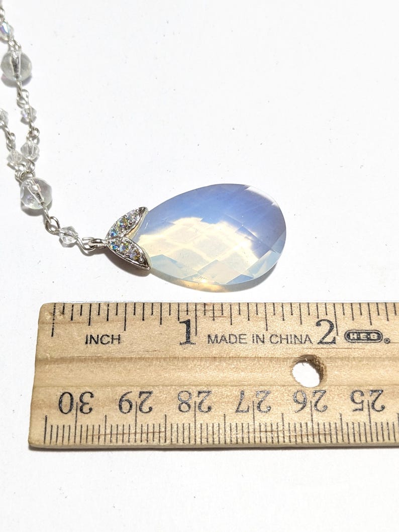 May include: A teardrop-shaped pendant necklace with a large, opalescent stone. The pendant is approximately 1 inch long and is attached to a delicate silver chain with small, clear beads. The pendant has a silver accent with small, clear stones.