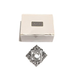 May include: A silver-toned brooch with the letter "C" inside a square frame with ornate detailing. The brooch is displayed next to a white box with the "AVON" logo.