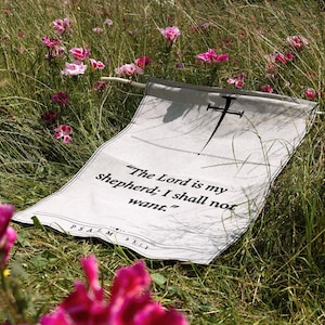 Puede incluir: Un tapiz de tela blanca colgado de una varilla de madera, con un gráfico negro de un clavo en la parte superior y el texto impreso "The Lord is my shepherd; I shall not want." seguido de "PSALM 23:1" en la parte inferior, expuesto al aire libre.