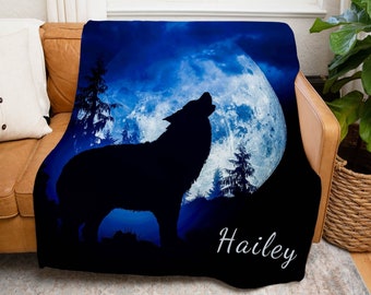 Puede incluir: Una manta de franela azul y negra con la silueta de un lobo aullando a la luna. La luna es un círculo grande, blanco brillante con una superficie texturizada. La manta tiene el nombre "Hailey" impreso en letras blancas cursivas en la esquina inferior derecha.