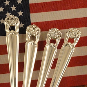 May include: Four silver spoons with ornate handles are displayed in a brown and white floral patterned container. The spoons are arranged in a fan shape with the handles pointing upwards. An American flag is in the background.