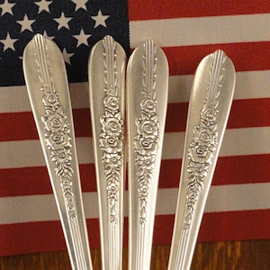 May include: Four silver-plated teaspoons with ornate floral designs. The spoons are arranged in a can with a floral pattern.