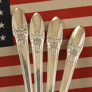 May include: Four silver spoons with ornate designs on the handles are arranged in a brown and white floral patterned cup. The spoons are positioned in a fan shape with the handles extending out of the cup.