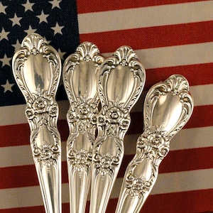 May include: Four silver-plated spoons with ornate handles are displayed in a floral patterned ceramic container. The spoons are arranged in a fan shape, with the handles pointing upwards. The container is sitting on a wooden surface.