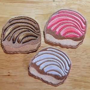 May include: Three embroidered patches shaped like a pastry with a swirl design. The patches are brown with a pink, white, and gold swirl design.