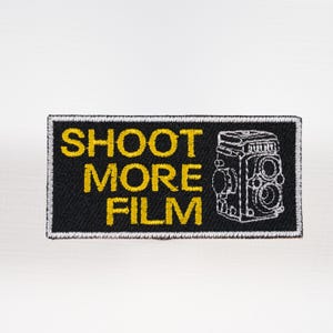 May include: Black rectangular patch with the words "SHOOT MORE FILM" in yellow, along with a white outline drawing of a vintage camera. The patch has a white border.