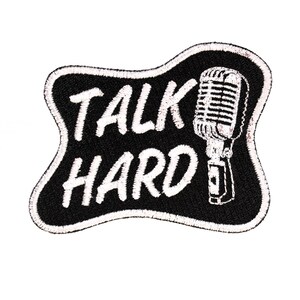 May include: An embroidered patch with a black background and a white border. The patch features the white text TALK HARD and a white illustration of a vintage-style microphone on the right side. The shape of the patch is irregular and wavy.