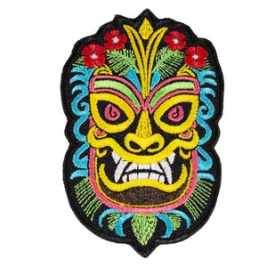 May include: An embroidered patch featuring a colorful tiki mask design on a black background. The mask has yellow skin, pink accents, white teeth, and blue swirling patterns. Red flowers and green leaves are stitched at the top and bottom edges.