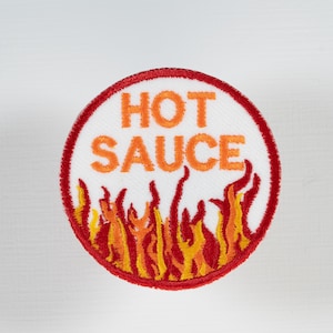 May include: Embroidered patch with a white background and a red circular border. The words "HOT SAUCE" are in orange block letters. Below the text is a stylized flame design in shades of red, orange, and yellow.