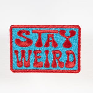 May include: A rectangular embroidered patch featuring a bright blue background with a thick red border. The text STAY WEIRD is stitched in a bold, red, stylized font across the center of the patch, filling the blue space with its distinct lettering.