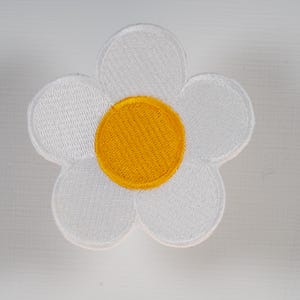May include: A five-petaled flower patch featuring white embroidered petals and a circular golden-yellow center. The design is stitched with a dense thread pattern, creating a textured surface with a defined border around the entire floral shape.