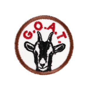 May include: A circular embroidered patch featuring a white background with a brown stitched border. In the center is a black and white goat head illustration. Above the goat head, the text G.O.A.T. is embroidered in red capital letters.
