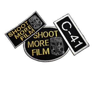 May include: Three embroidered patches with black backgrounds and white borders. Two patches feature yellow text reading SHOOT MORE FILM and a camera icon. The third rectangular patch features white text reading C-41 in a vertical orientation.