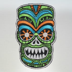 May include: Embroidered patch featuring a stylized tiki mask design. The mask has a green, blue, orange, and black color scheme with intricate details. The eyes are circular with starburst patterns, and the mouth displays sharp teeth.