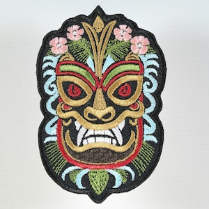 May include: Embroidered patch featuring a colorful tiki mask design. The mask has red, green, and gold accents, with pink flowers and blue swirls. The patch is outlined in black.