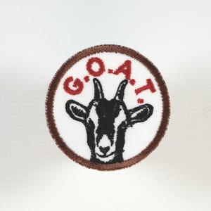May include: Circular patch with a goat illustration in black and white, surrounded by a brown border. The word "G.O.A.T." is written in red above the goat's head. The patch is on a white background.