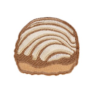 May include: A round embroidered patch shaped like a concha sweet bread. The design features a light brown base with a textured, cream-colored top layer arranged in a curved, shell-like pattern with distinct stitched lines throughout the surface.