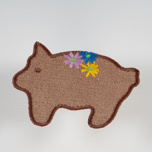 May include: A brown pig-shaped embroidered patch with a dark brown stitched border. The design features three small embroidered flowers on the pig's back, including one light purple flower, one blue flower, and one yellow flower with green centers.