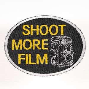 May include: An oval-shaped embroidered patch with a black background and a white border. The patch features the yellow text SHOOT MORE FILM stacked on the left side and a white outline illustration of a twin-lens reflex camera on the right.