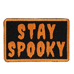 May include: A rectangular embroidered patch with a black background and an orange border. The text STAY SPOOKY is embroidered in orange, bold, sans-serif capital letters across the center of the patch, arranged in two lines of text.