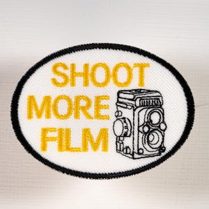 May include: An oval-shaped embroidered patch with a white background and a thick black border. The text SHOOT MORE FILM is stitched in yellow thread. To the right of the text is a black-stitched outline of a twin-lens reflex camera.