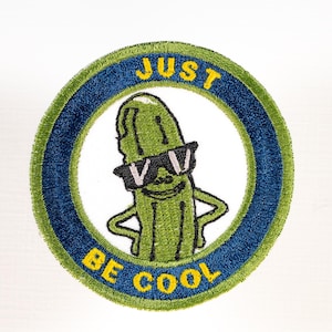 May include: A circular embroidered patch featuring a green pickle character wearing black sunglasses. The design is set against a white background with a dark blue border and a lime green outer edge. The yellow text reads JUST BE COOL.
