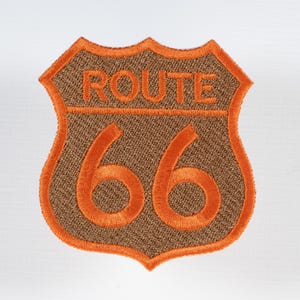 May include: Embroidered Route 66 patch in a shield shape. The patch features the words "ROUTE" above the number "66" in orange thread. The background is a textured brown fabric, with an orange border.