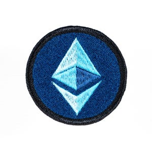 May include: A circular embroidered patch featuring a dark blue background with a central light blue diamond-shaped geometric symbol. The patch is finished with a thick black stitched border that frames the entire circular design clearly.