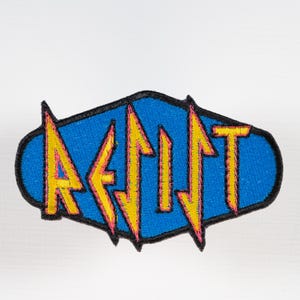 May include: Embroidered patch featuring the word "AEJIT" in yellow, outlined in pink, against a blue background. The patch has a black border and an oval shape. The design has a retro, vintage aesthetic.