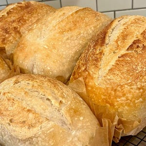 May include: Several loaves of freshly baked bread with golden-brown crusts are arranged on a metal rack. The bread is wrapped in parchment paper and has a rustic appearance, with visible scoring on the top.