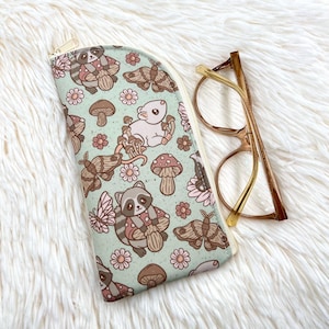 May include: A soft-sided glasses case with a whimsical woodland pattern featuring raccoons, mushrooms, butterflies, and flowers in shades of brown, pink, and green. The case is next to a pair of gold-rimmed eyeglasses.