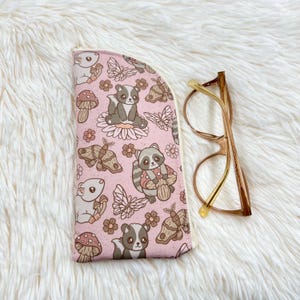 May include: A pink glasses case with a woodland animal pattern, including raccoons, butterflies, and mushrooms. The case is next to a pair of gold-rimmed eyeglasses.