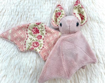 Könnte beinhalten: Ein Fledermaus-Plüschtier aus weichem, hellrosa Stoff. Die Innenseiten der Ohren und die Unterseiten der Flügel bestehen aus cremefarbenem Stoff mit einem Muster aus rosa Rosen und kleinen weißen Blumenakzenten. Zwei kleine schwarze Augen.