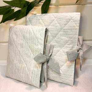 May include: Two quilted fabric cases with a white and light green dotted pattern. The cases are tied with gray bows. The cases are in different sizes, and the larger one is open. The background is white.