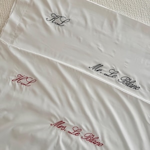 Op de afbeelding: Witte lakens met geborduurde letters. Eén laken heeft "KL" in zwart garen. Een ander laken heeft "Mr. Le Blanc" in zwart garen. Een derde laken heeft "KL" en "Mrs. Le Blanc" in rood garen.