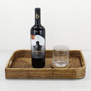 May include: A rectangular brown woven tray with handles holds a bottle of red wine and a clear glass. The wine bottle has a black label with white text and a boat illustration. The glass sits on a small coaster.