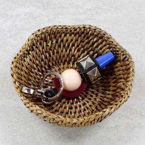 May include: A small, woven rattan basket holds a collection of items. Inside, there's a set of keys, a light pink sphere, a dark red disc, and a black and silver object with a blue top. The basket is a natural brown color.