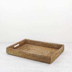May include: A rectangular woven rattan tray in a natural brown color. The tray has a raised edge and cut-out handles on each end. The woven pattern is visible throughout the tray's surface.