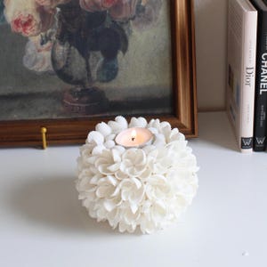May include: A spherical candle holder covered in small, white, overlapping seashells arranged in a floral pattern. A single lit tea light candle sits in the center of the top. The structure is round and textured with numerous individual shells.