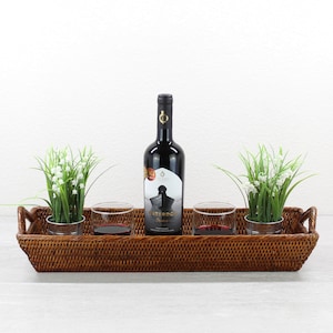 May include: A rectangular, woven rattan serving tray holds a bottle of red wine, two glasses with red wine, and two small potted plants with white flowers. The tray has handles on each end.