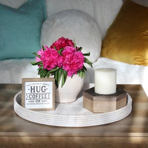 May include: A white woven tray holds a vase of pink peonies, a white candle, and a wooden box. A small framed sign reads "Can't decide if I need a hug, coffee, or an entire week of sleep."