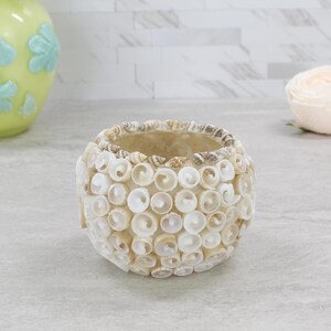 May include: A round candle holder covered in small, white, spiral-shaped seashells. The exterior is densely packed with these shells, creating a textured, spherical surface. The rim is lined with a row of smaller, tan and brown patterned shells.