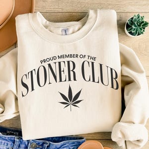 Può includere: Una felpa beige con il testo "Proud Member of the Stoner Club" e una grafica di foglia di marijuana nera.