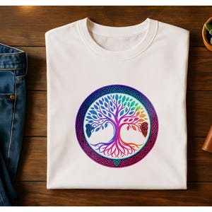 May include: White t-shirt featuring a colorful tree of life design within a circular frame. The tree's leaves and branches display a gradient of colors, including blue, green, and red. The design is set against a wooden surface, with blue jeans and a small plant visible.